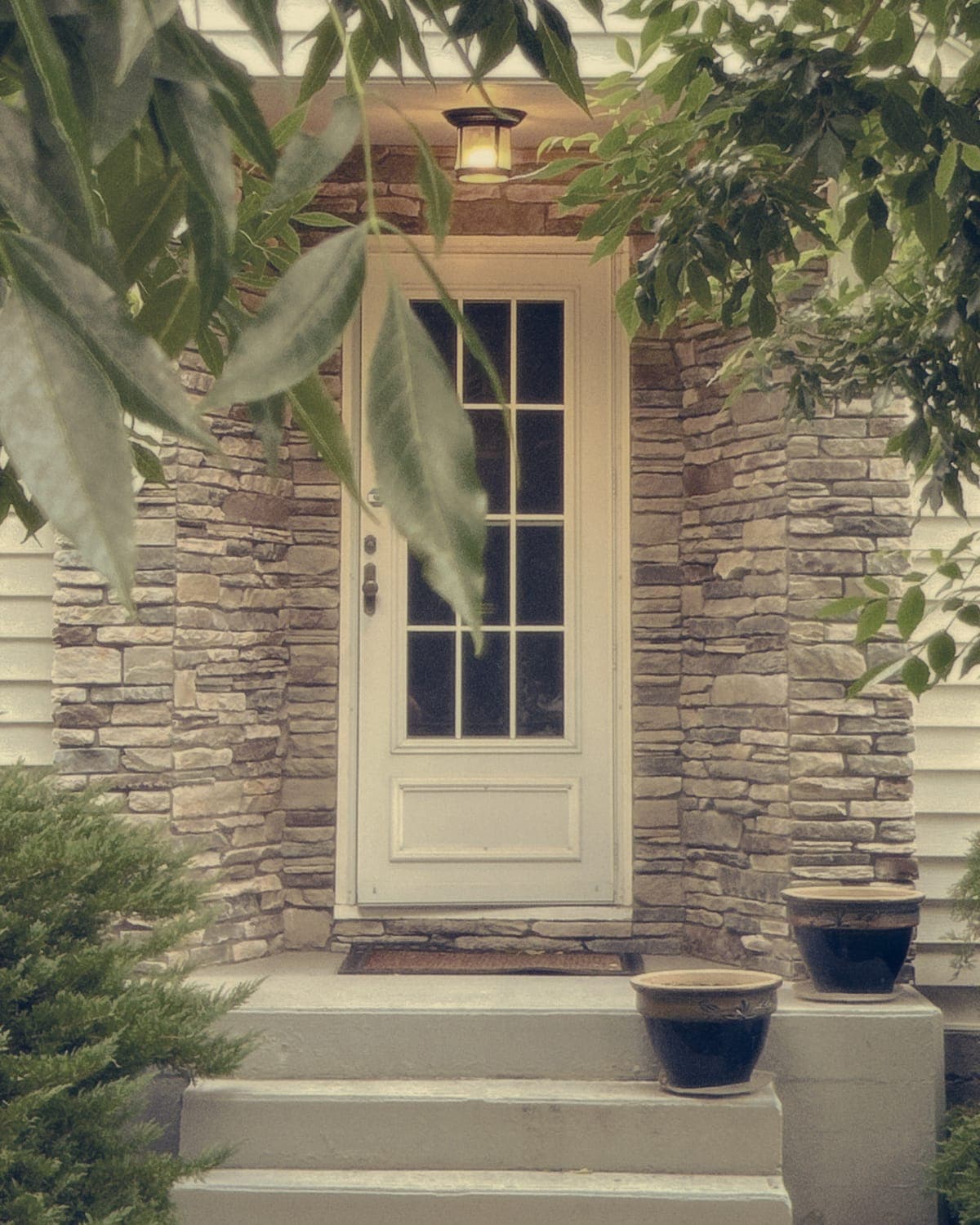 A white front door in a stone porch, with a lit lantern above and leafy branches framing the steps